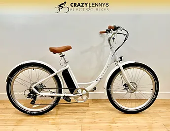 A white electric bicycle is displayed indoors against a light-colored wall with a wooden floor, giving it a stylish home setting. The bicycle features brown handlebars and a brown saddle, with the logo "Crazy Lenny's Electric Bikes" visible on the wall behind it.