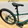 A green Reid E-Trail 2.0 MY23 electric bike is shown from the side, focusing on the rear half. The bike has a thick, spiked rear tire, a visible battery compartment integrated into the frame, and a comfortable black seat. The background features a wooden floor and a white wall. A green Reid E-Trail 2.0 MY23 electric bike is shown from the side, focusing on the rear half. The bike has a thick, spiked rear tire, a visible battery compartment integrated into the frame, and a comfortable black seat. The background features a wooden floor and a white wall.