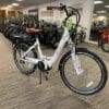 A white electric bicycle with a brown leather saddle, the EG Athens 350MD Mid Drive, is displayed prominently in a well-lit bike shop. Numerous other bicycles are lined up neatly in the background, showcasing a wide variety of styles and colors. The floor is carpeted with a striped pattern. A white electric bicycle with a brown leather saddle, the EG Athens 350MD Mid Drive, is displayed prominently in a well-lit bike shop. Numerous other bicycles are lined up neatly in the background, showcasing a wide variety of styles and colors. The floor is carpeted with a striped pattern.