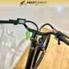 A black electric bike, Reid Quest 1.0 MY23, with a digital display on the handlebars is shown resting against a white wall on a wooden floor. The bike has a green tag attached near the handlebars. The background features the logo and text "Crazy Lenny's Electric Bikes. A black electric bike, Reid Quest 1.0 MY23, with a digital display on the handlebars is shown resting against a white wall on a wooden floor. The bike has a green tag attached near the handlebars. The background features the logo and text "Crazy Lenny's Electric Bikes.