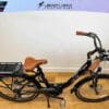 A black EG Athens 350MD Mid Drive electric bike with a brown leather saddle and handles is displayed indoors on a wooden floor. It features a rear cargo rack, mounted battery, mid drive 350MD motor, and front suspension. The wall in the background has a logo that reads "Crazy Lenny's Electric Bikes. A black EG Athens 350MD Mid Drive electric bike with a brown leather saddle and handles is displayed indoors on a wooden floor. It features a rear cargo rack, mounted battery, mid drive 350MD motor, and front suspension. The wall in the background has a logo that reads "Crazy Lenny's Electric Bikes.