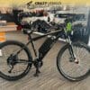 A black electric bicycle is displayed in a showroom with a banner above reading "Crazy Lenny's Electric Bikes." The bike features a robust frame, wide tires, and a central electric motor. Various biking accessories and products are visible in the background, highlighting an EG Kyoto (Demo). A black electric bicycle is displayed in a showroom with a banner above reading "Crazy Lenny's Electric Bikes." The bike features a robust frame, wide tires, and a central electric motor. Various biking accessories and products are visible in the background, highlighting an EG Kyoto (Demo).