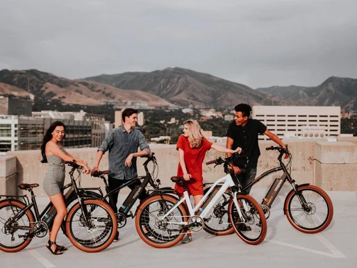 Four people standing with ebikes on a rooftop with mountains in the background. They appear to be engaged in conversation and smiling. The sky is overcast, and the group looks relaxed and happy, perhaps discussing the latest ebike regulations.