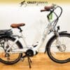A white EG Athens 350MD Mid Drive electric bicycle with a brown seat and black handlebars is displayed in a store. The bike features a rear-mounted battery, a kickstand, front and rear fenders, and a mounted headlight. A sign above reads "Crazy Lenny's Electric Bikes. A white EG Athens 350MD Mid Drive electric bicycle with a brown seat and black handlebars is displayed in a store. The bike features a rear-mounted battery, a kickstand, front and rear fenders, and a mounted headlight. A sign above reads "Crazy Lenny's Electric Bikes.