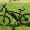 A black electric bike sits on green grass under a sunny sky. The EG Kyoto (Demo) has a thick frame with an integrated battery, front suspension, and disc brakes. Above the bike, the text "Crazy Lenny's Electric Bikes" is displayed along with a stylized bike logo, perfect for an exciting demo ride. A black electric bike sits on green grass under a sunny sky. The EG Kyoto (Demo) has a thick frame with an integrated battery, front suspension, and disc brakes. Above the bike, the text "Crazy Lenny's Electric Bikes" is displayed along with a stylized bike logo, perfect for an exciting demo ride.