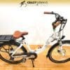 A white electric bike with brown handlebars and seat is displayed on a wooden floor. The EG Athens 350DX Hub Drive model has a battery and rear rack, showcasing "Crazy Lenny's Electric Bikes" at the top center. Featuring a hub drive motor, front suspension fork, and digital display, the bike exudes modern sophistication.