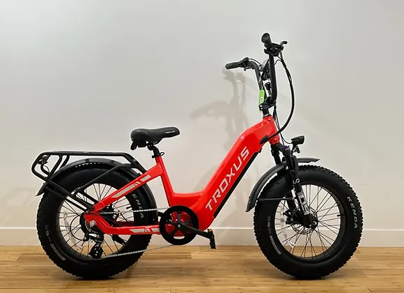 A bright red Troxus Lynx 20'' Fat Tire E-bike with wide black tires is displayed against a plain white wall on a wooden floor. The fat tire e-bike features a sturdy frame, a back rack, and an upright handlebar design.