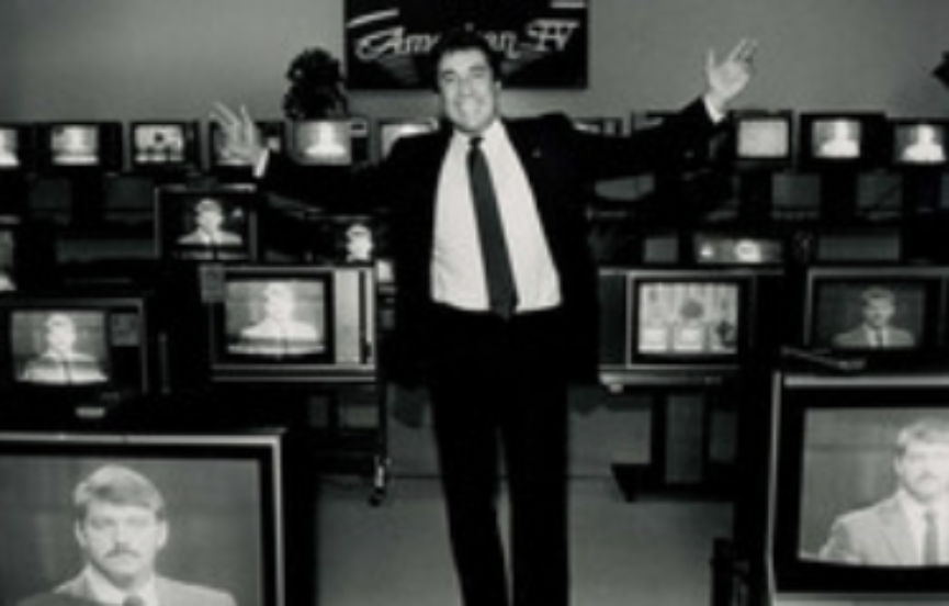 A man in a suit stands with arms raised in front of multiple televisions displaying different images of various individuals. The background boasts a sign reading "American TV." The setting appears to be an electronics store showroom, perhaps introducing the "About Us" section. The image is in black and white.