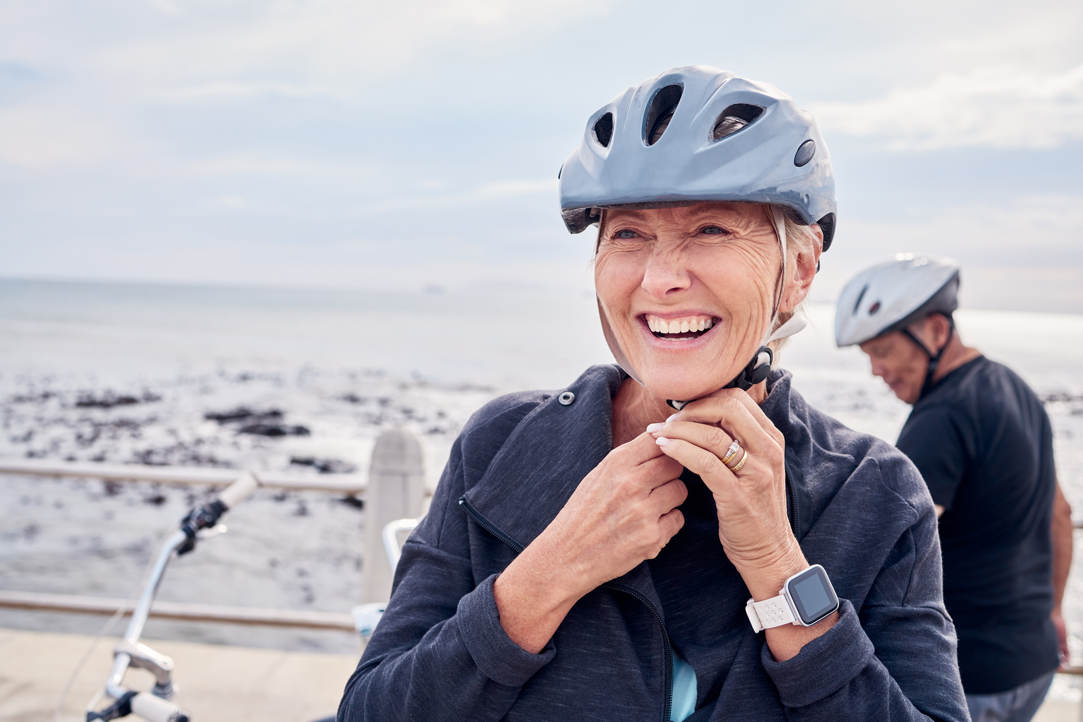 Smiling woman adjusts bike helmet by the sea.