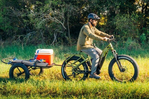 Person biking with trailer carrying cooler in field.