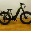 A green fat tire electric bicycle with a rear cargo rack stands on a wooden floor against a plain white wall. The bike features thick tires, a central battery, and front suspension. Green fat tire electric bike on wooden floor