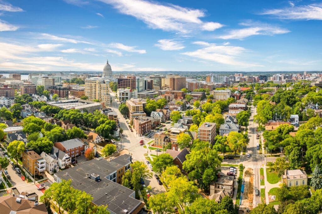 Aerial view of a city with a prominent white-domed capitol building, tree-lined streets, and a mix of residential and commercial buildings under a bright blue sky with scattered clouds.