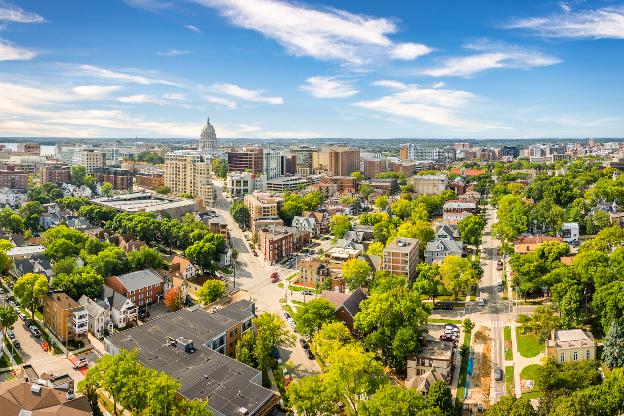 Aerial view of a city with a prominent white-domed capitol building, tree-lined streets, and a mix of residential and commercial buildings under a bright blue sky with scattered clouds.