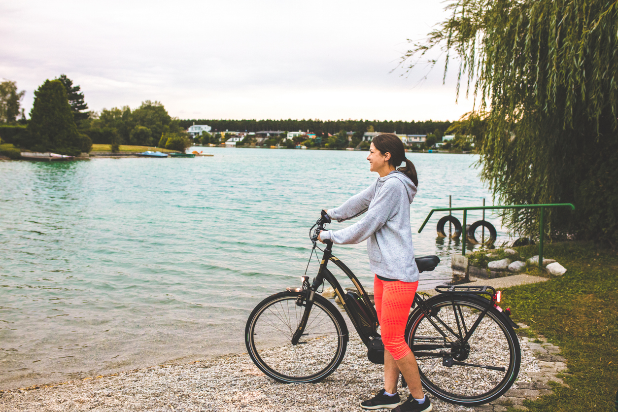 A woman in a gray hoodie and bright orange leggings stands beside her bicycle, looking out over a calm lake—maybe she’s just enjoyed an E Bike Test Ride from Crazy Lenny’s before taking in the peaceful view.