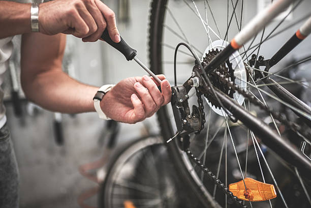 A person uses a screwdriver to adjust the rear derailleur on a bicycle. The close-up shows their hands working on the bike’s gear mechanism, with the bike wheel and gears clearly visible.