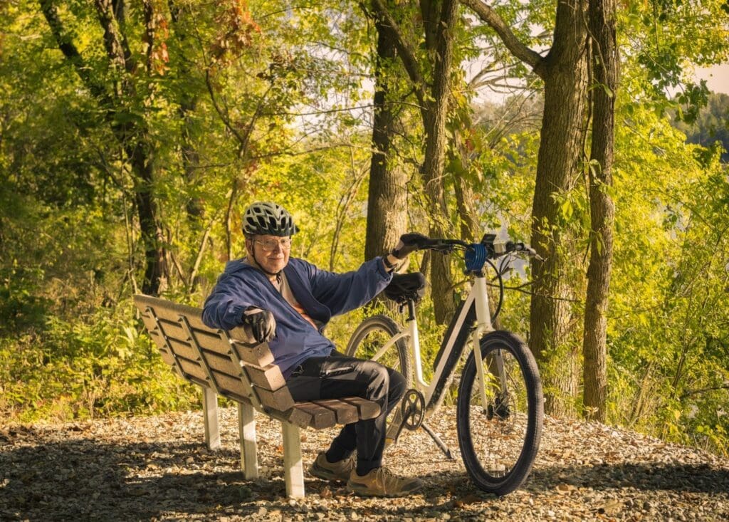 Elderly man resting on bench with bicycle in forest.